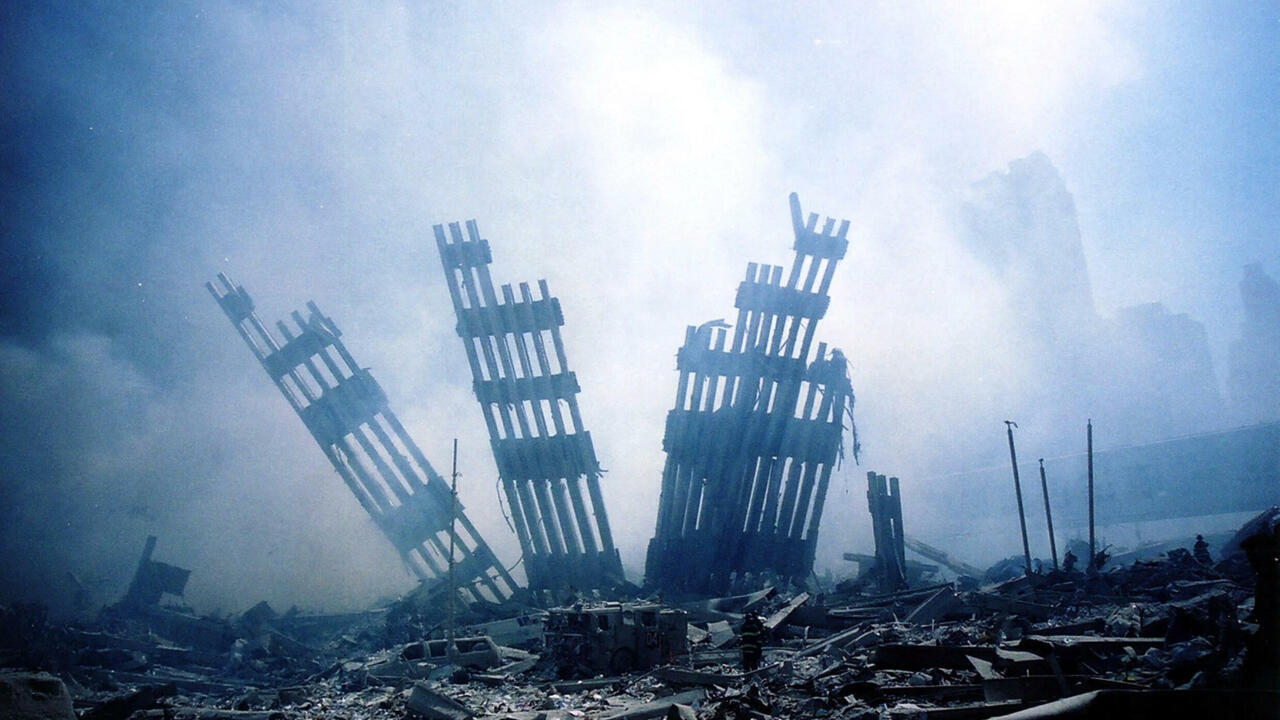 Smoke and dust surround damaged high-rise structures amid extensive rubble and emergency vehicles.