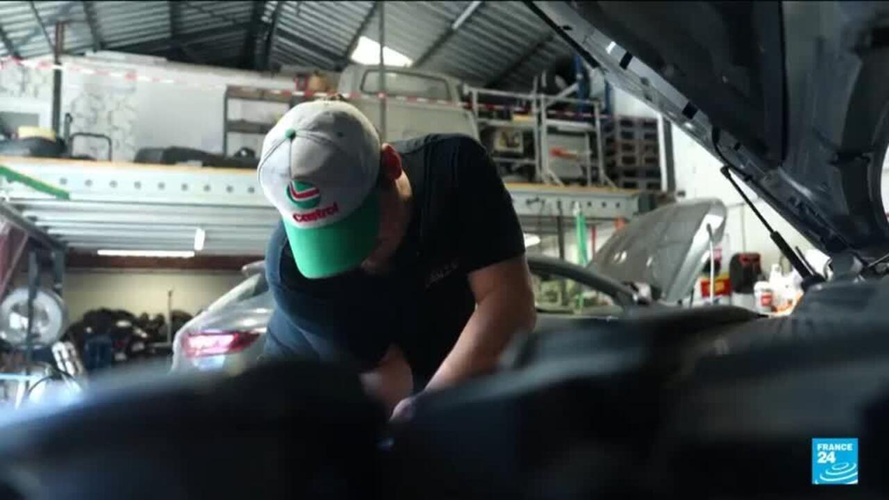 Mechanic in a green and white cap works under the hood of a car in a garage.