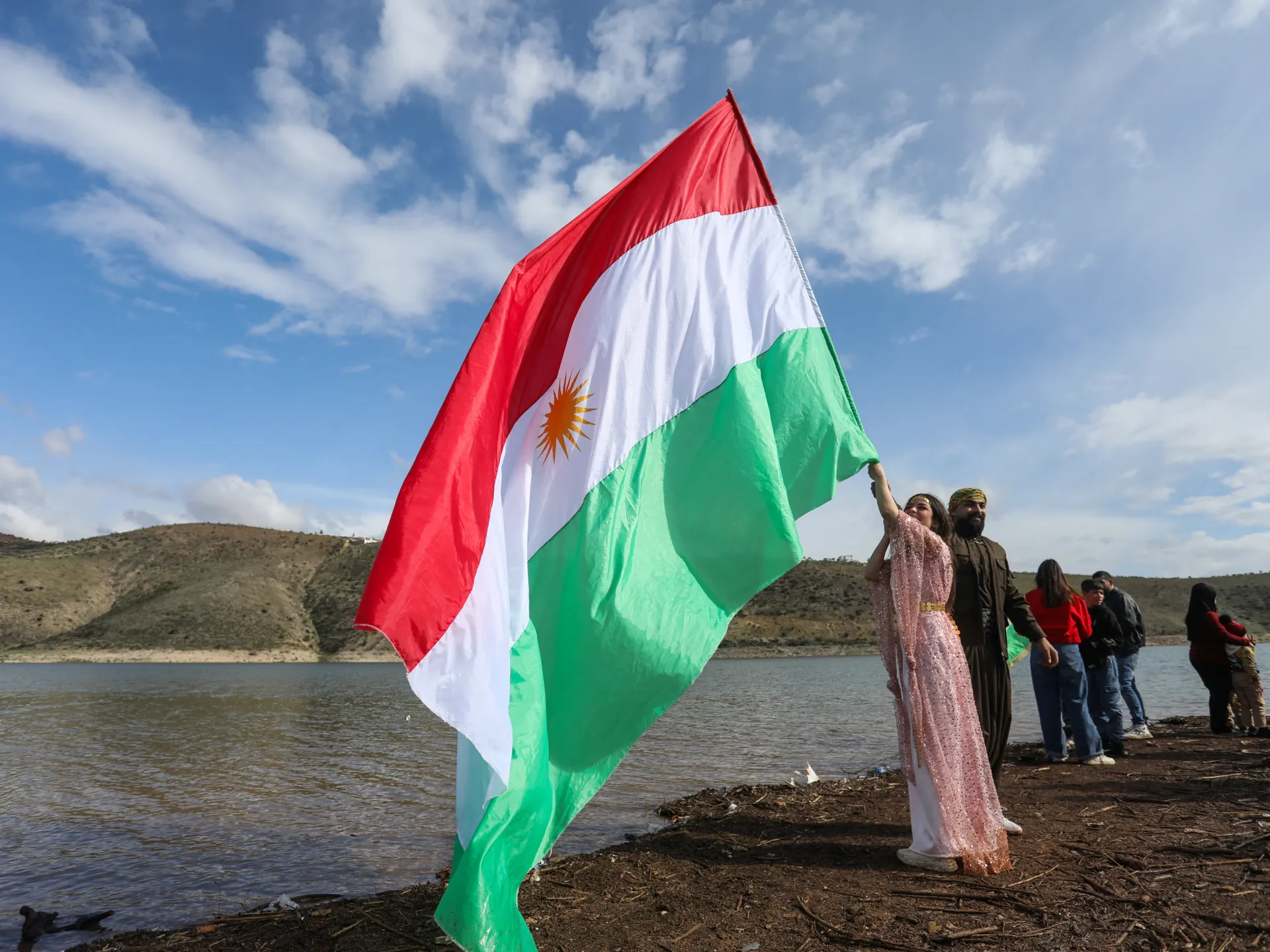 People hold a Kurdish flag beside a lake under a partly cloudy sky.