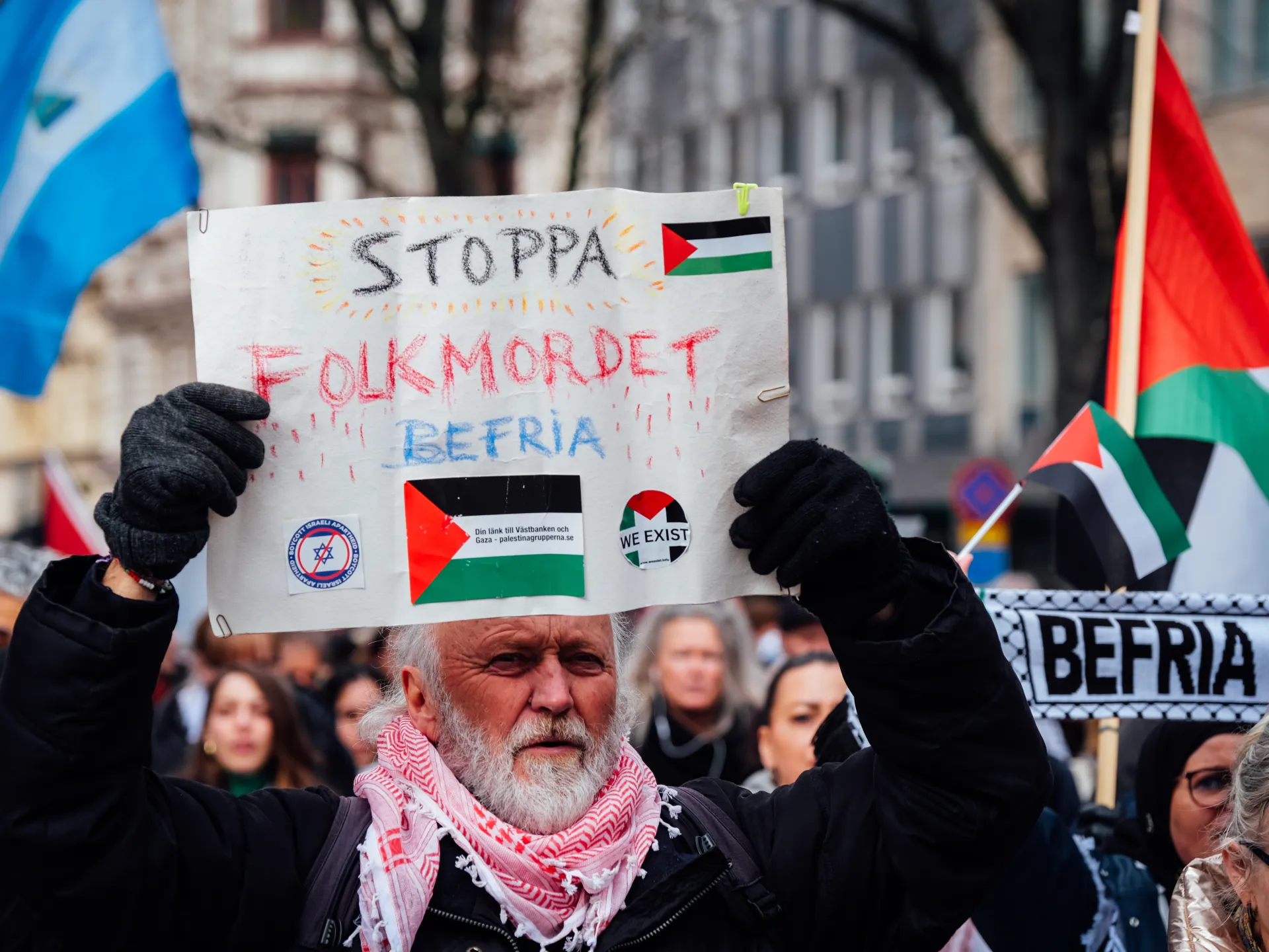 Protester holding a handwritten Swedish sign reading “Stoppa folkmordet” amid Palestinian flags at a demonstration.