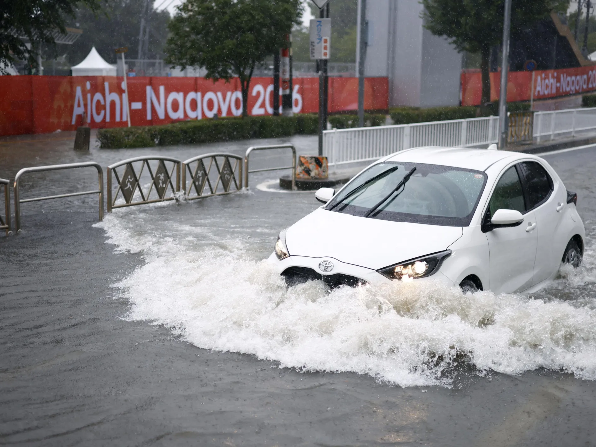 White car drives through deep floodwater on a rain-soaked Japanese city street.