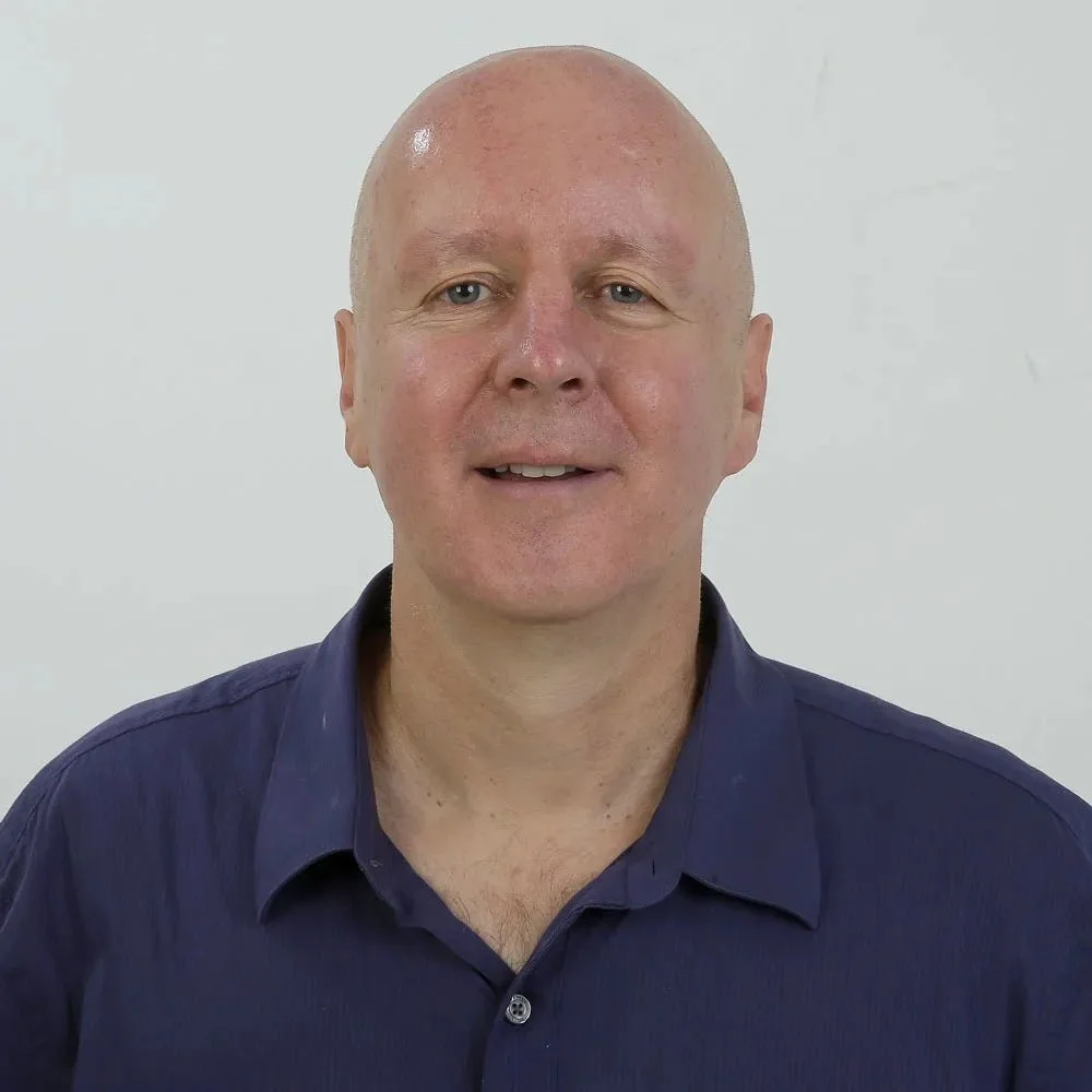 Bald man in a dark blue collared shirt facing the camera against a light background.