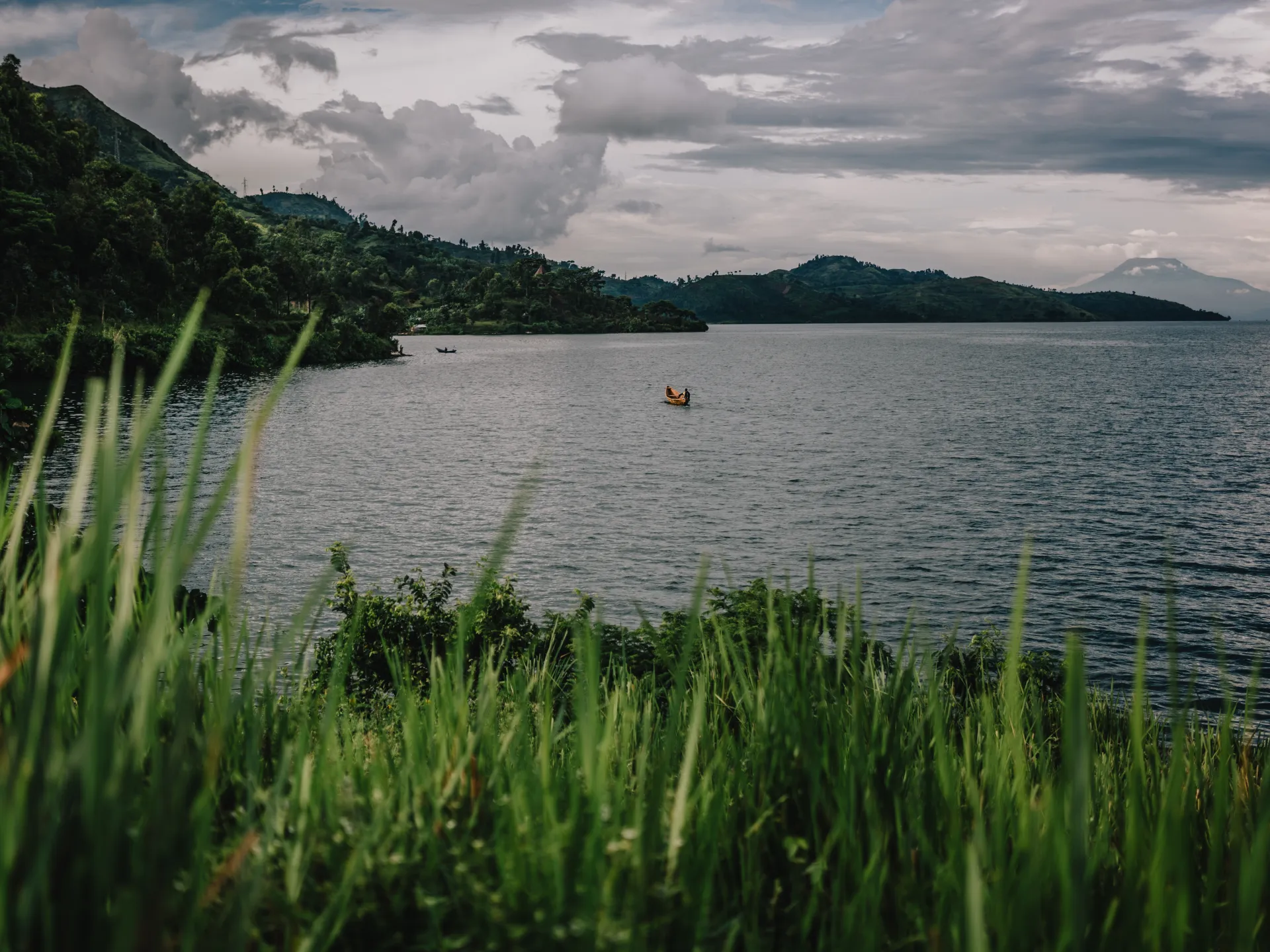 A small boat crosses a broad lake beneath cloudy skies, with green hills and tall grasses in the foreground.