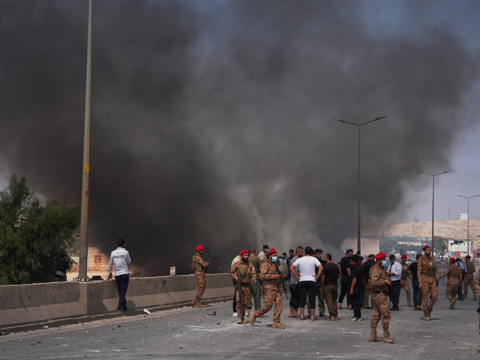 Smoke billows over a roadway as soldiers, police and civilians gather near a blast site.