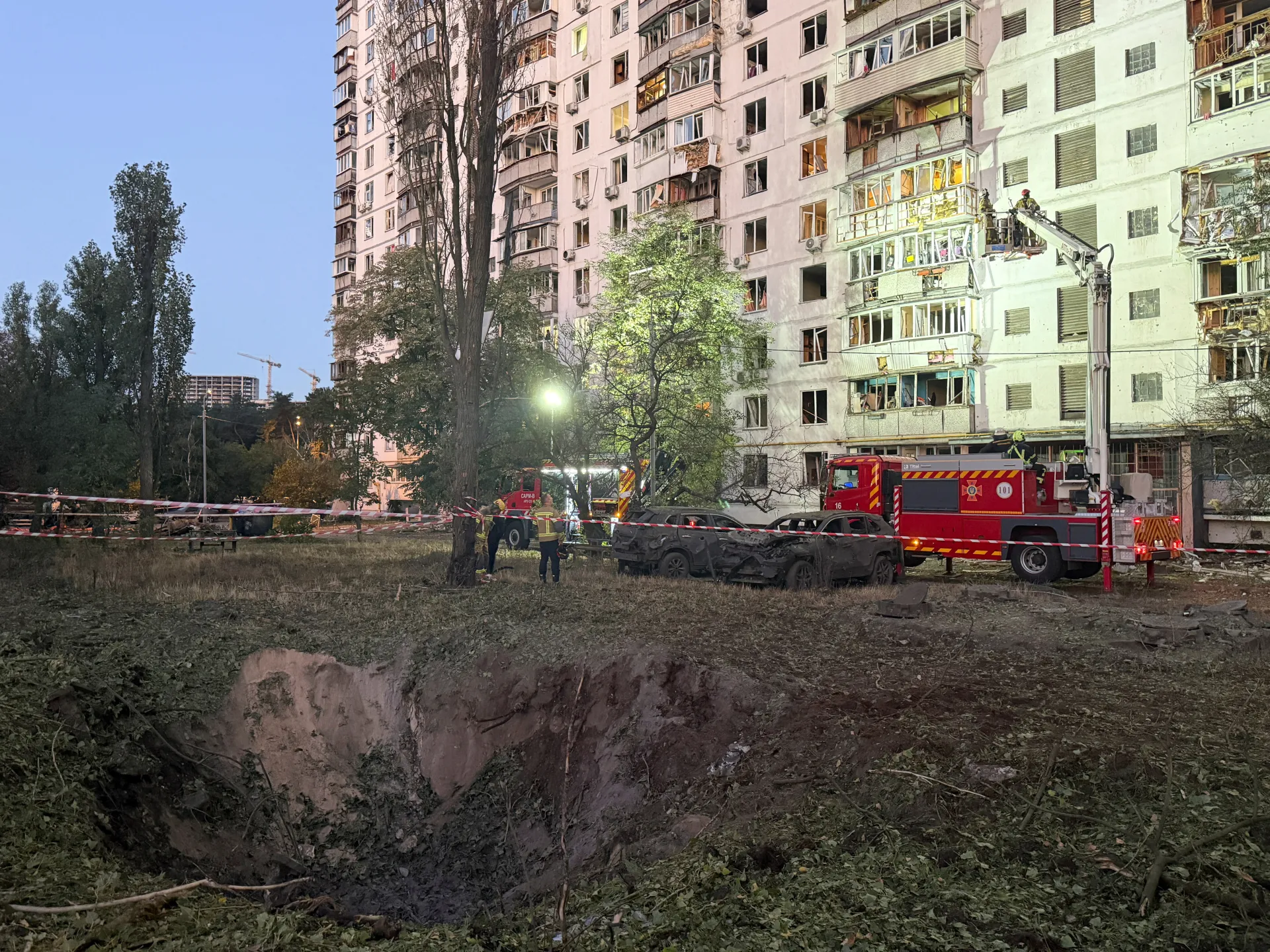 Firefighters and emergency vehicles outside a damaged apartment building beside a large blast crater