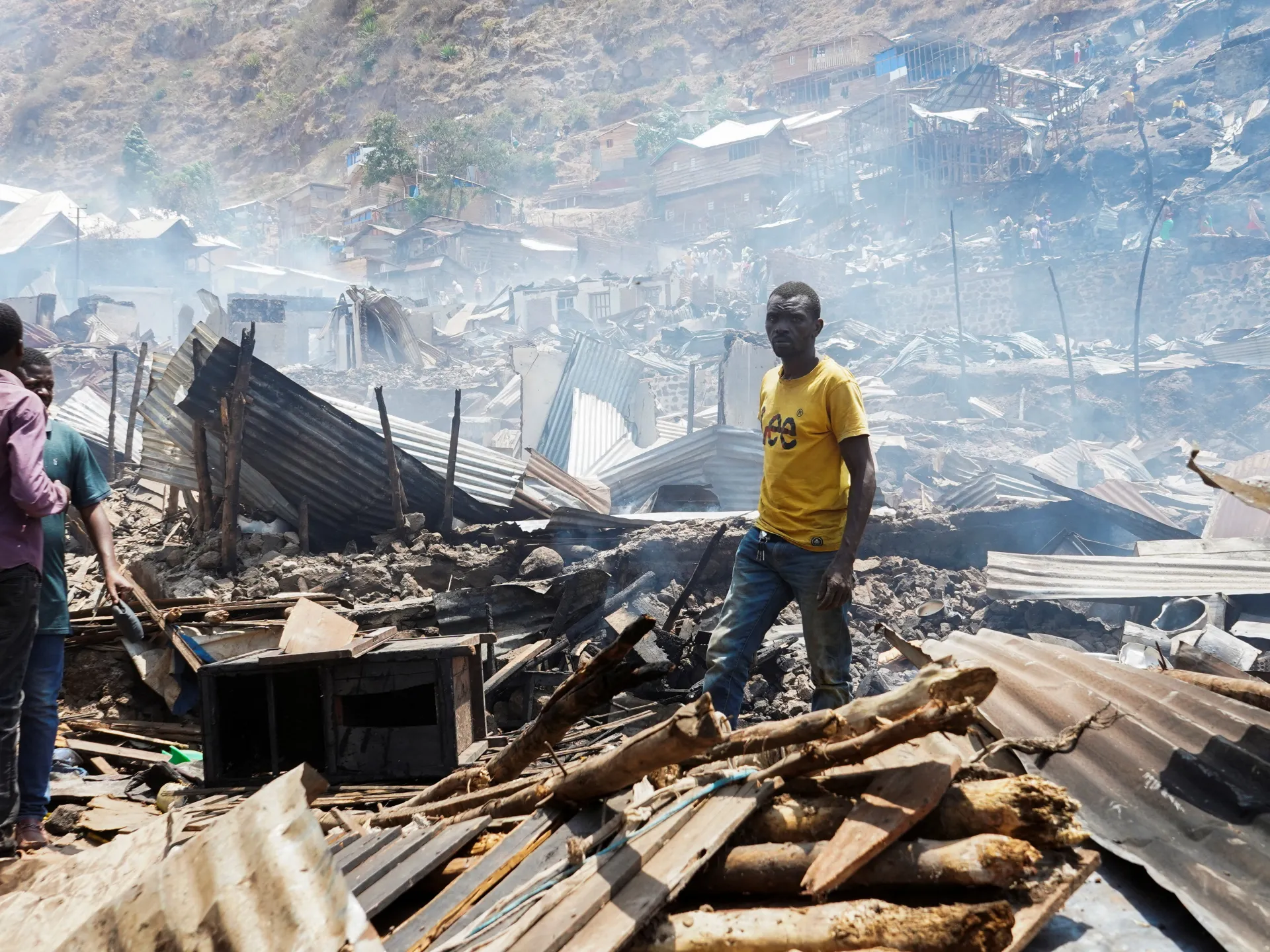 People stand amid extensive rubble and collapsed structures in a smoke-filled hillside settlement.