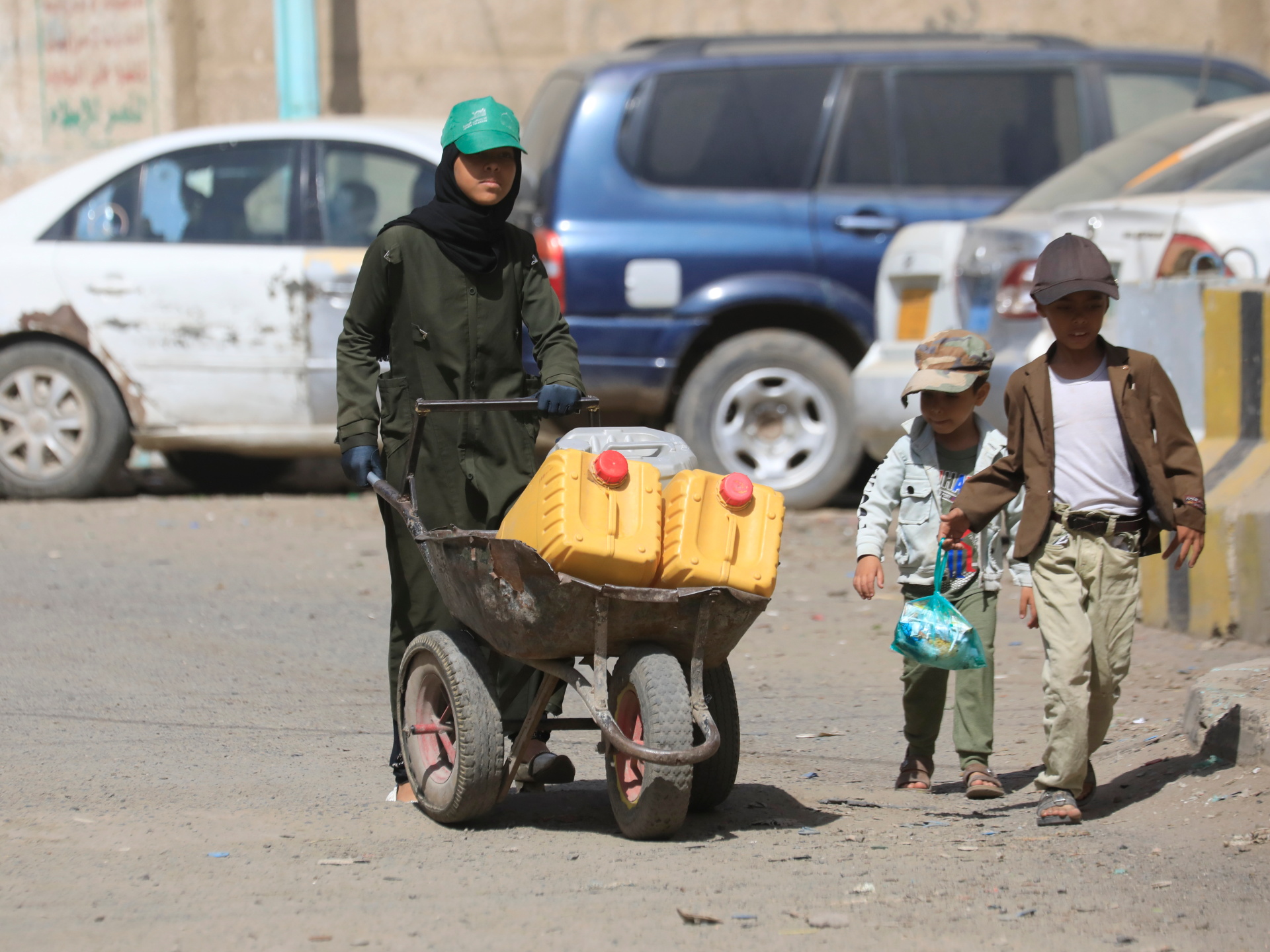 Woman pushes a wheelbarrow carrying yellow water containers beside two boys on a dusty street