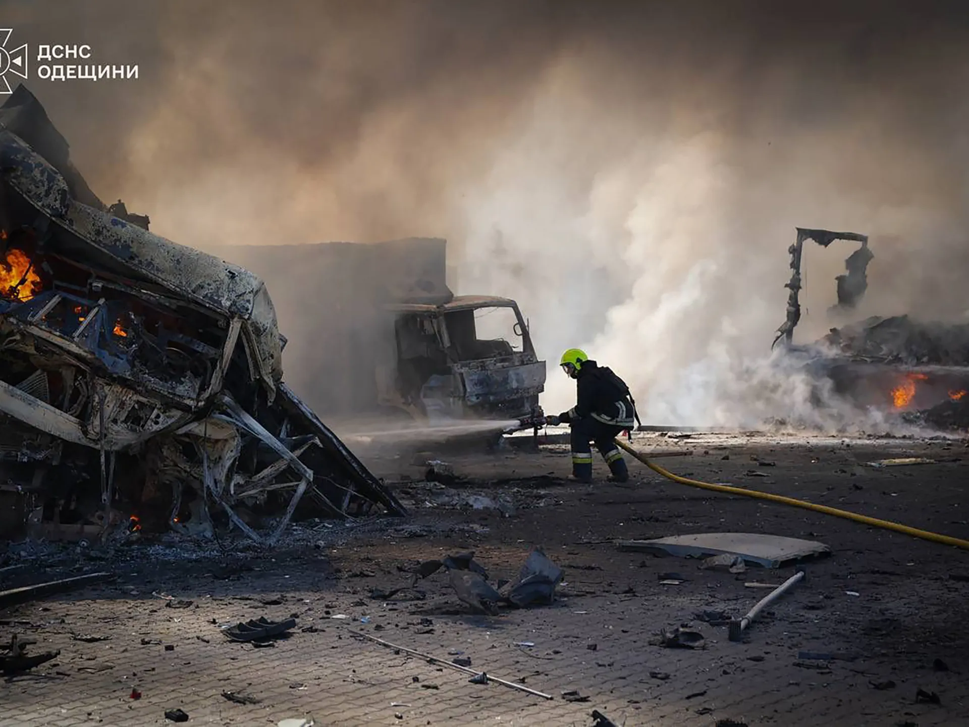 Firefighter sprays water amid burning wreckage, smoke, debris and damaged vehicles at a disaster scene.