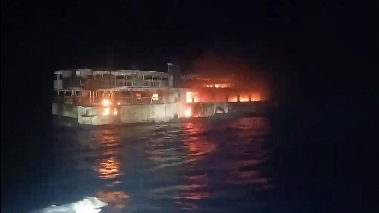 A ferry burns at night, with flames glowing through windows and reflecting on dark water.