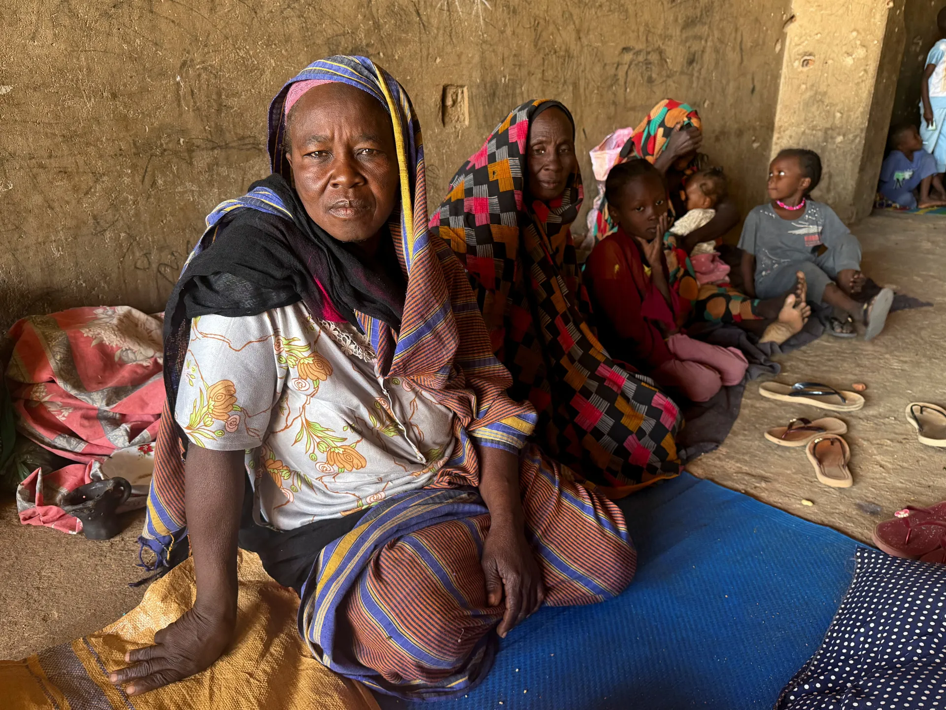 Two women and several children sit inside a modest mud-walled room.