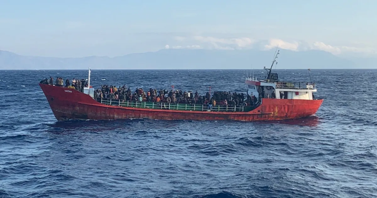 Red fishing vessel crowded with people sailing across rough open sea beneath distant mountains