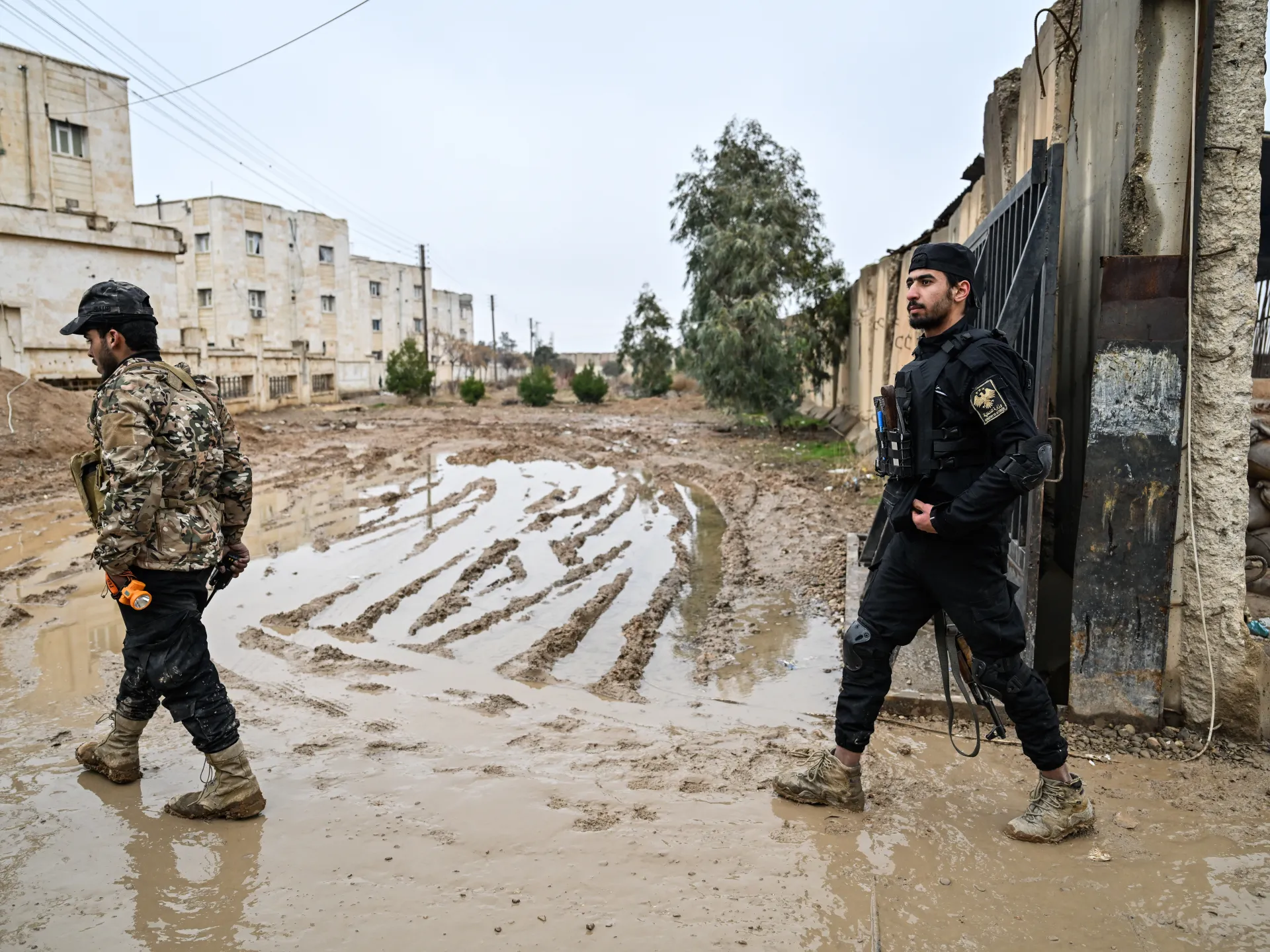 Two armed men walk through a muddy, flood-covered street beside damaged buildings.
