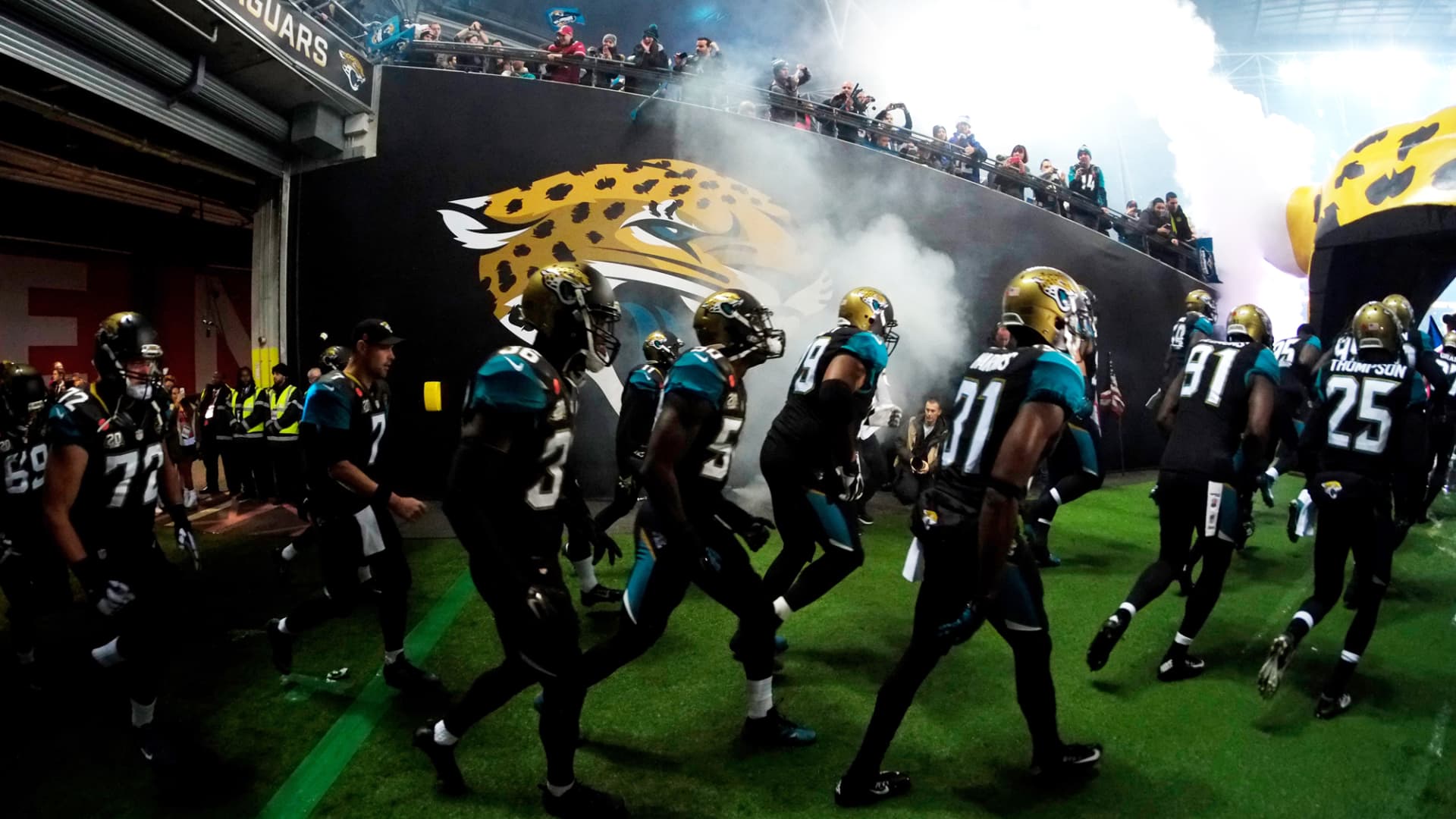 Jacksonville Jaguars players run onto the field through smoke beneath a large team logo and cheering spectators.