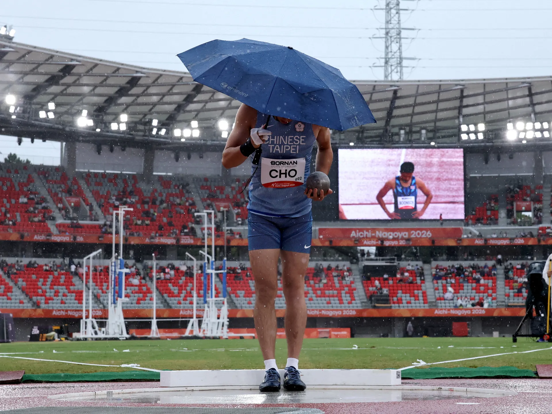 An athlete holds a shot put beneath a blue umbrella during rain in a stadium.