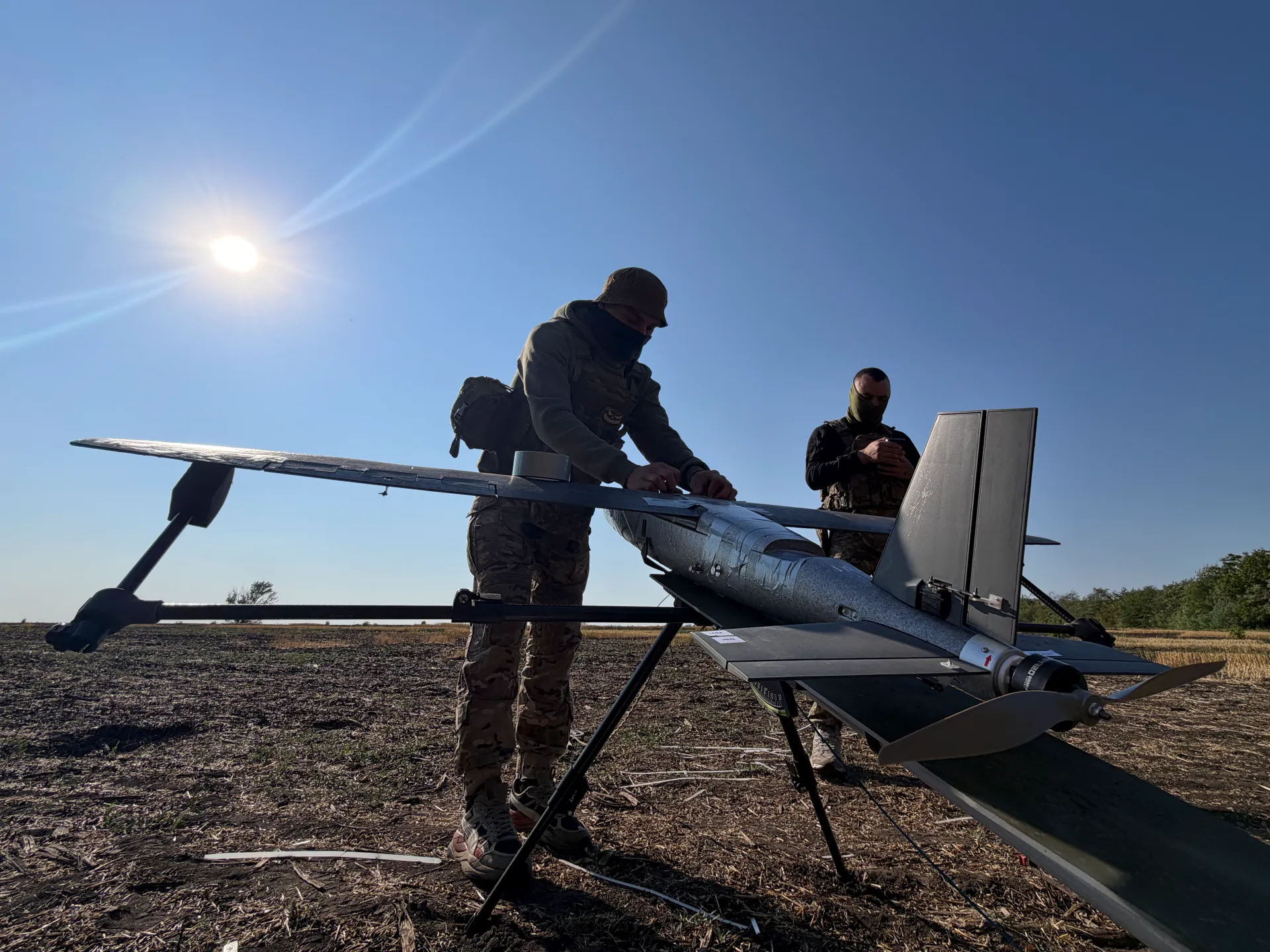 Two masked soldiers inspect a fixed-wing drone outdoors under bright sunlight.