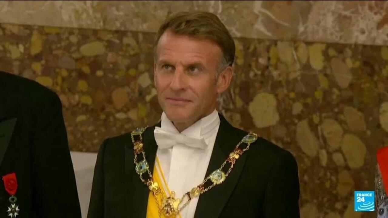 A man in a black formal suit, white bow tie, and ceremonial chain stands before a patterned stone wall.