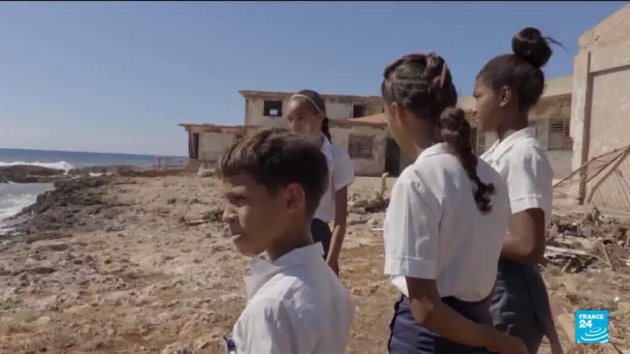 Children in white shirts stand on a rocky shoreline beside weathered seaside buildings.