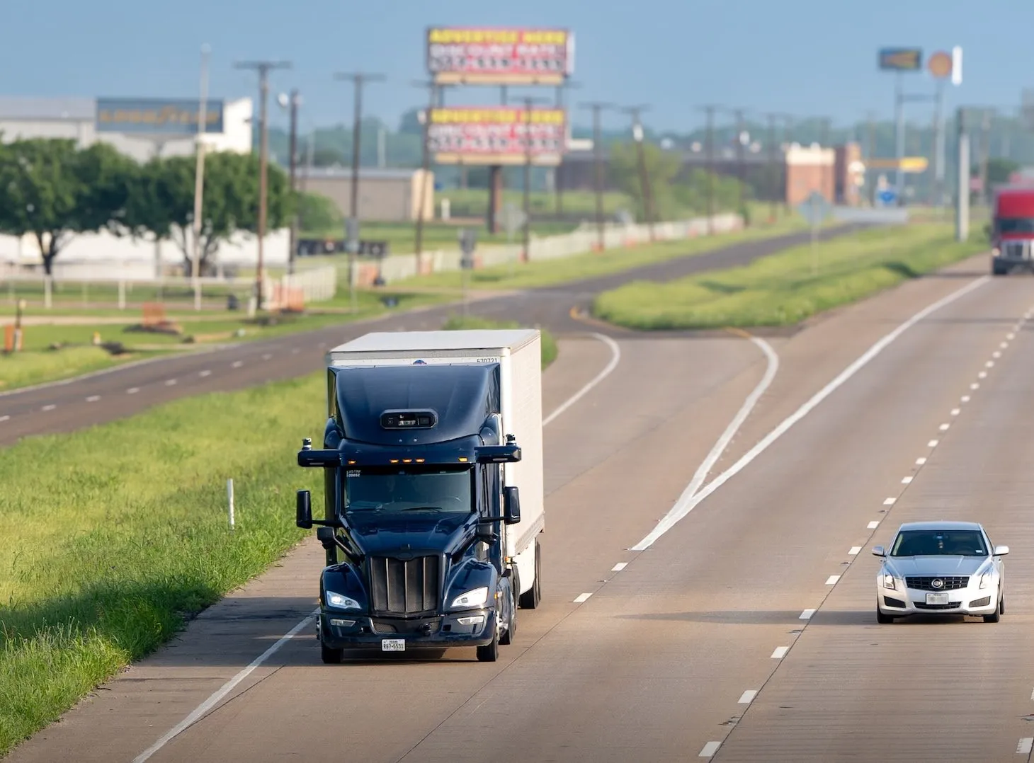 A dark semi truck and white sedan travel on a divided highway under clear skies.
