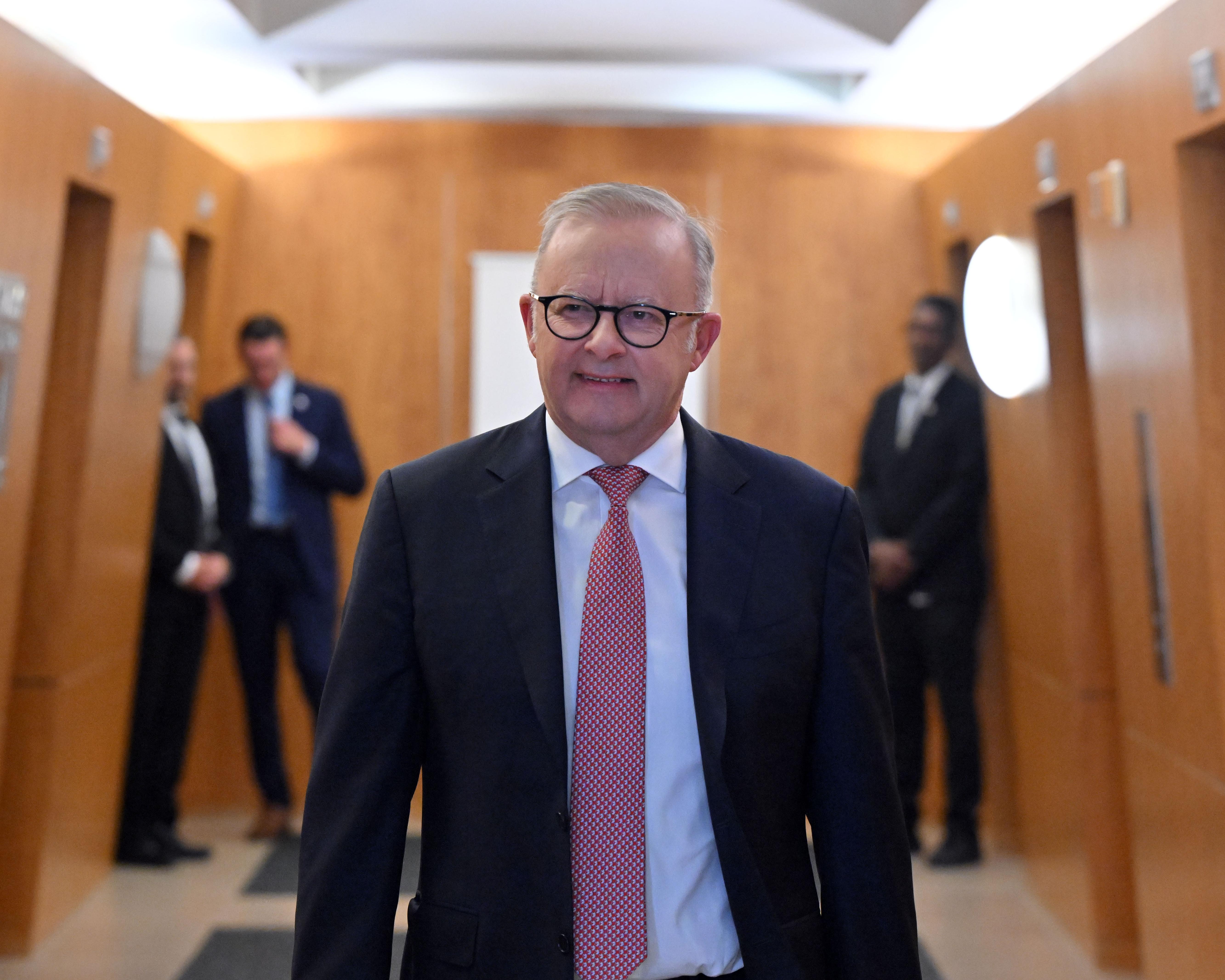 Grey-haired man in glasses and suit walks through a wood-paneled corridor with officials behind him.