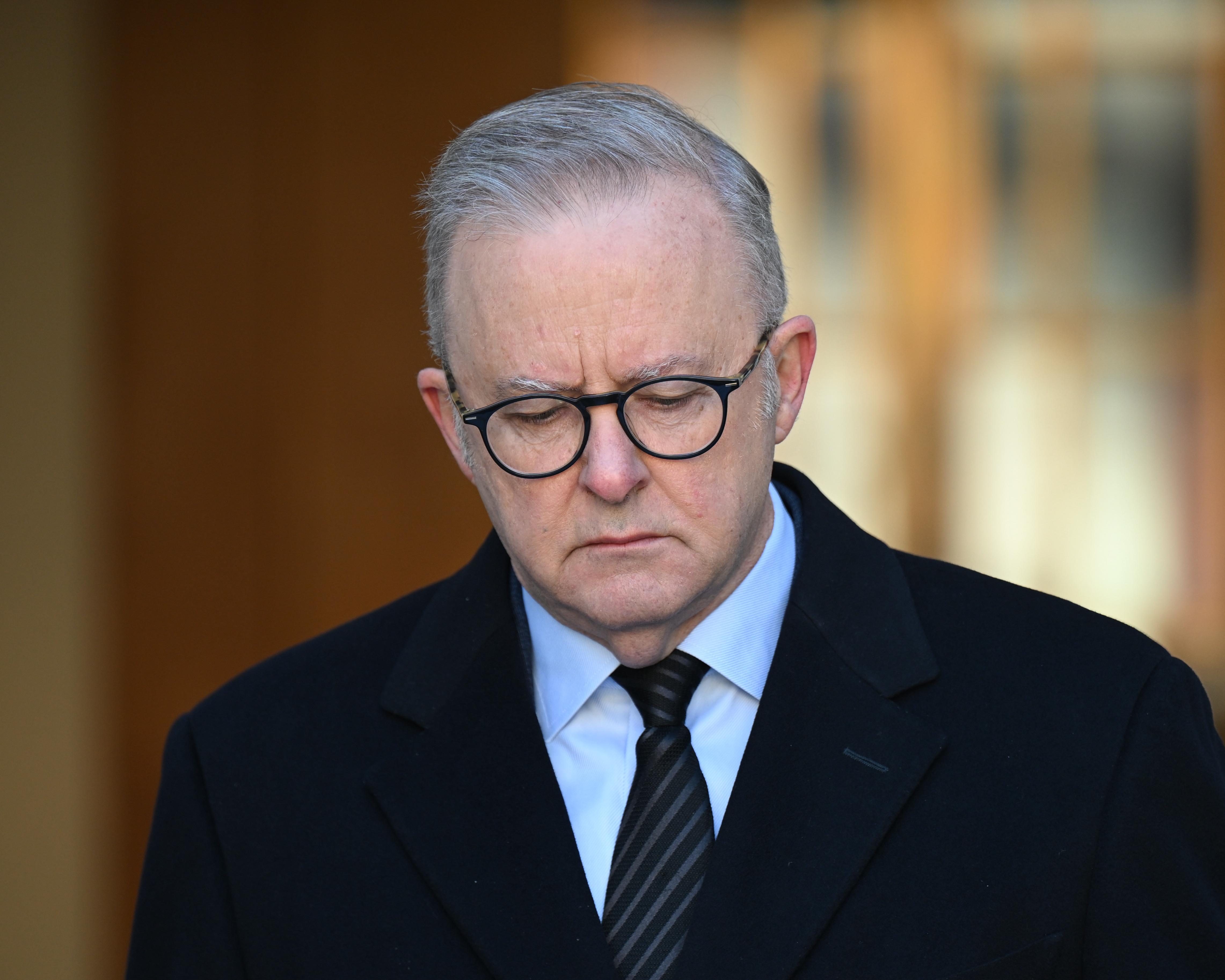 Gray-haired man wearing glasses, dark coat and striped tie, looking downward indoors.