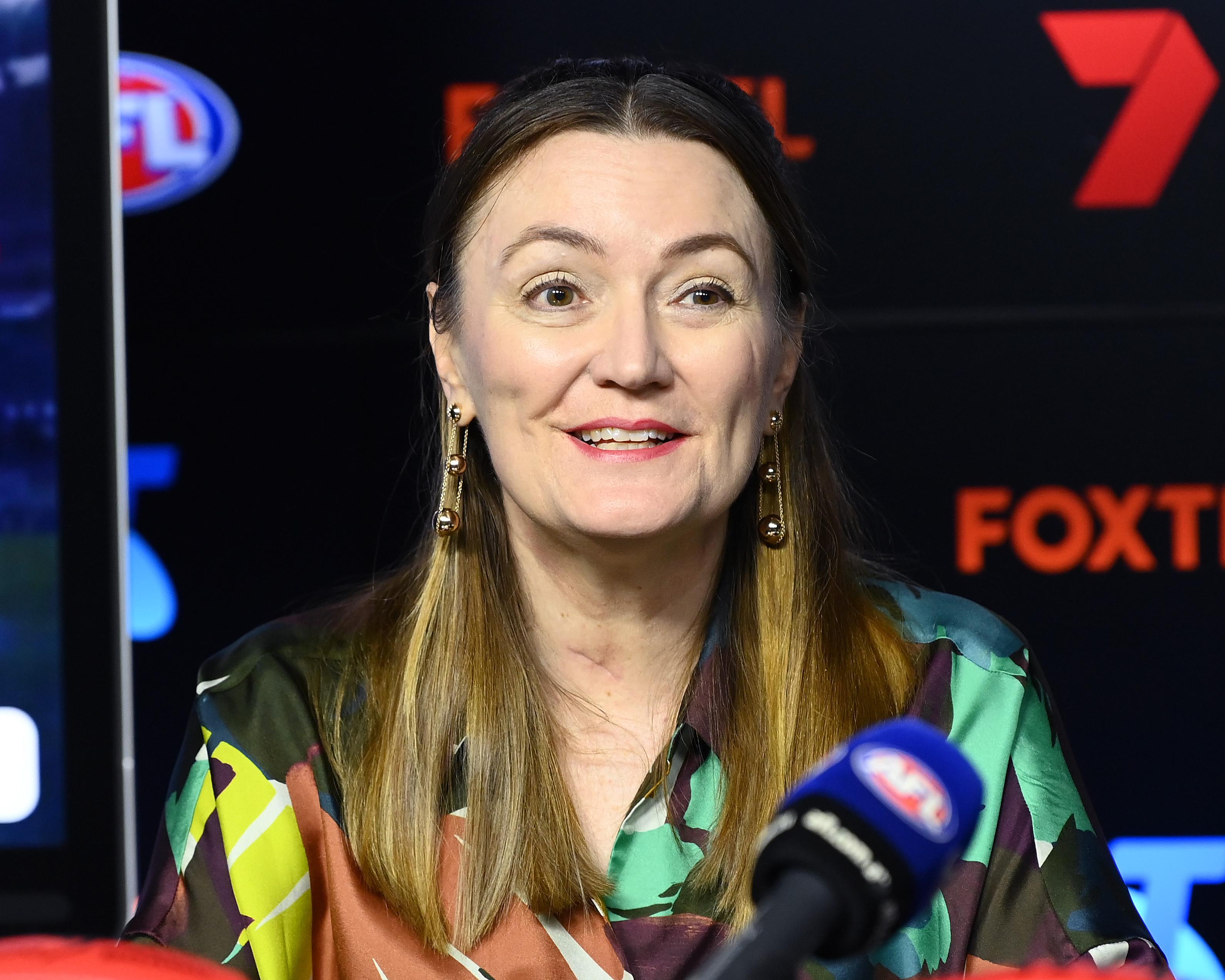 Woman speaking at a press event with microphones in front of a branded media backdrop