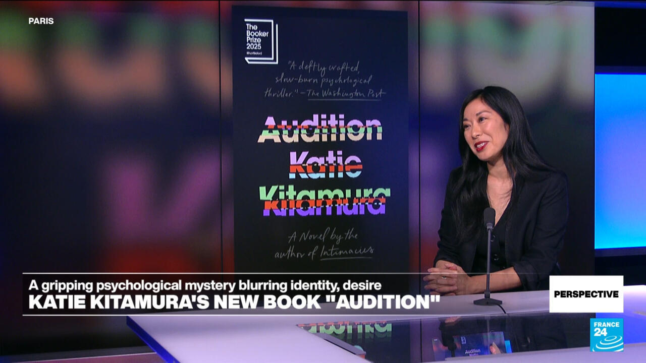 Katie Kitamura sits at a studio desk beside a large display of her book Audition.