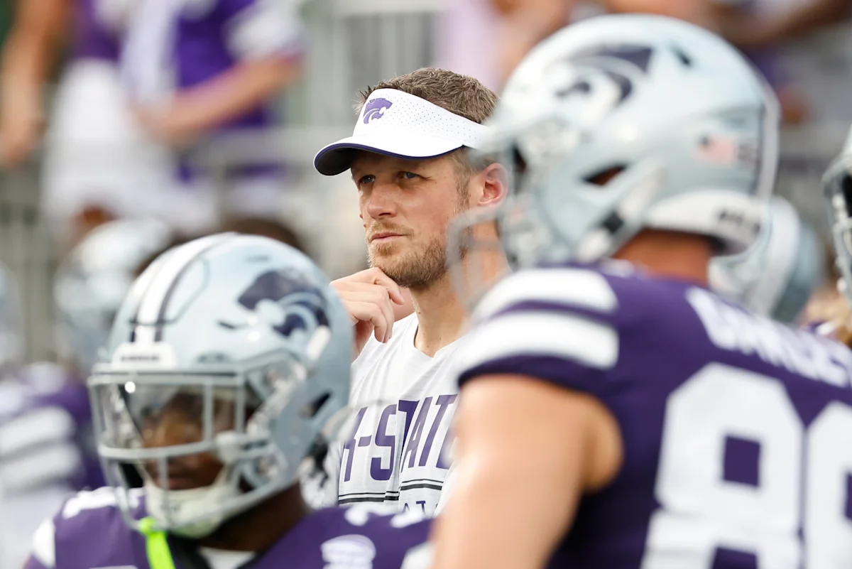 Kansas State football player in visor watches from the sideline amid teammates wearing helmets.