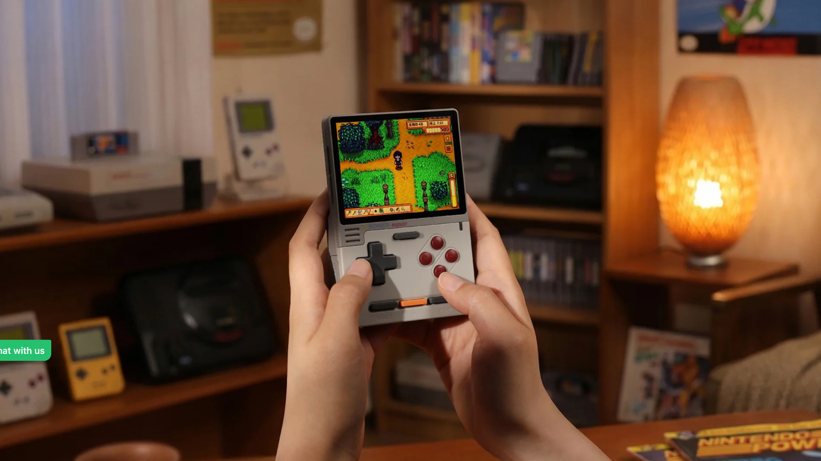 Hands hold a gray handheld gaming device displaying a colorful pixel art game in a room with retro consoles.