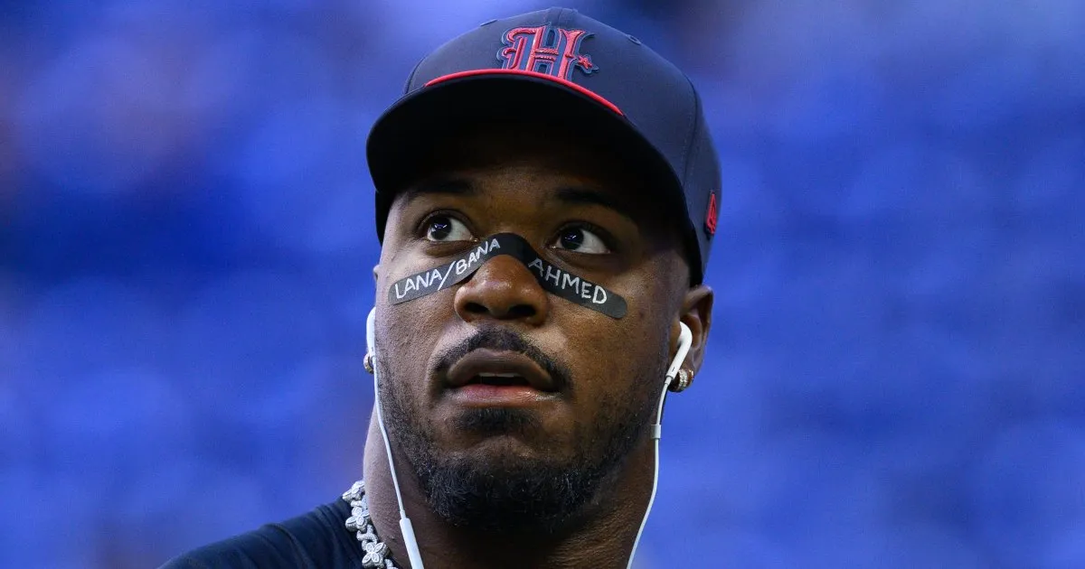 A football player wears a navy cap, eye black, earrings, and white earbuds against a blue background.