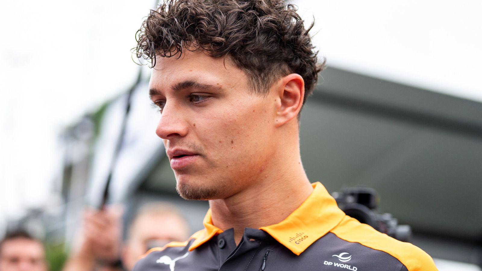 Young man with curly dark hair wearing a yellow-collared McLaren team shirt, shown in profile.