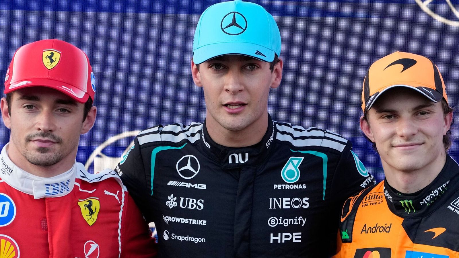 Three Formula 1 drivers pose together in team racing suits and caps at a podium event.