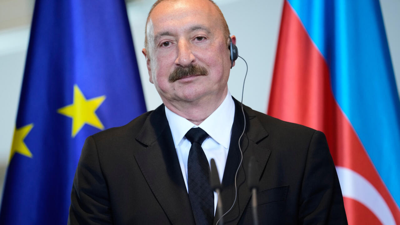 A mustached man in a dark suit wears an earpiece, with Azerbaijan and French flags behind him.