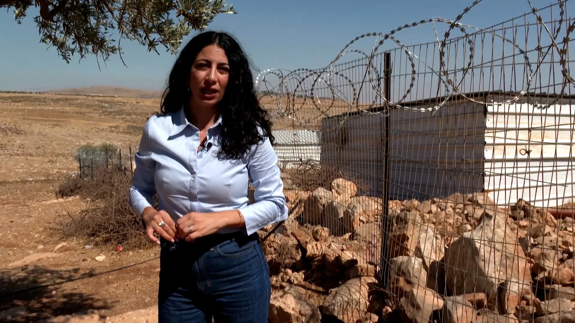 Woman standing beside a barbed-wire fence in a dry, rocky landscape under clear skies