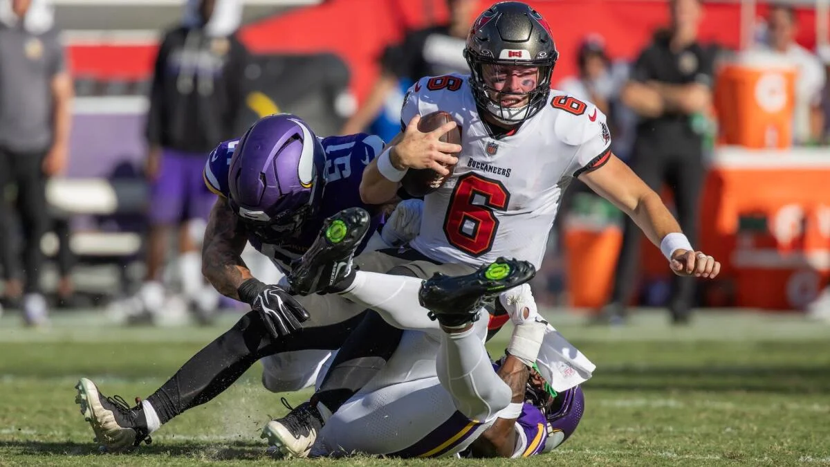 Buccaneers quarterback in a white No. 6 jersey is tackled by a Vikings defender in purple.
