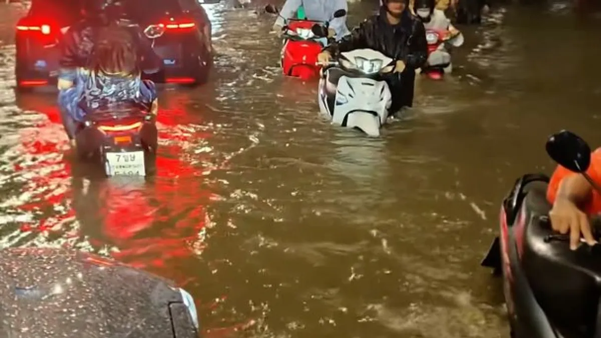 People maneuver scooters through deep floodwater beside cars on a rain-flooded urban road at night.