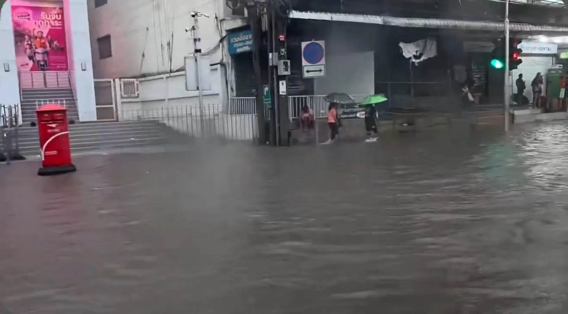 Floodwater covers a city street as pedestrians shelter beneath umbrellas near storefronts and traffic lights.