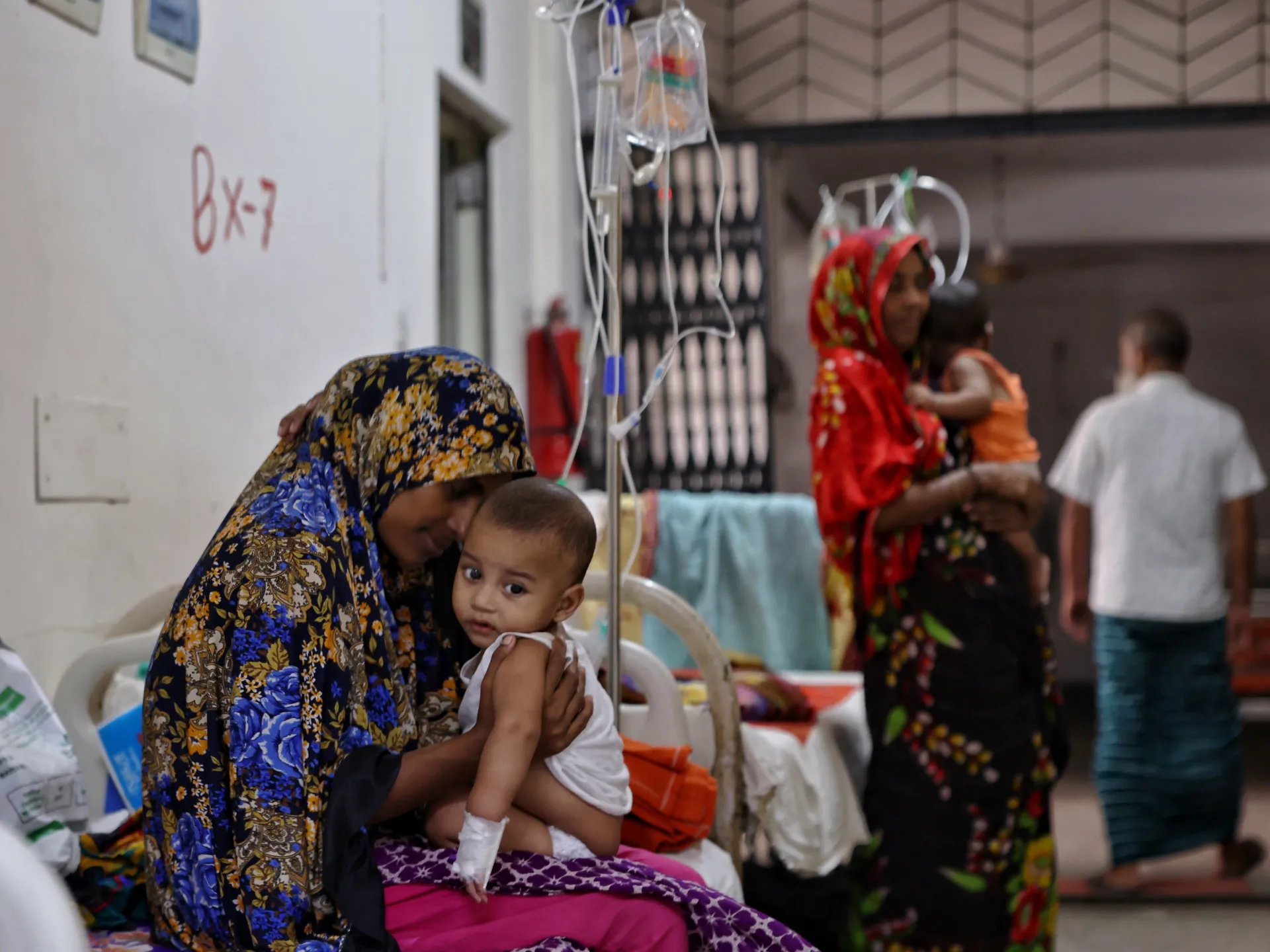 Woman in patterned headscarf holds an infant receiving intravenous treatment inside a crowded hospital ward.