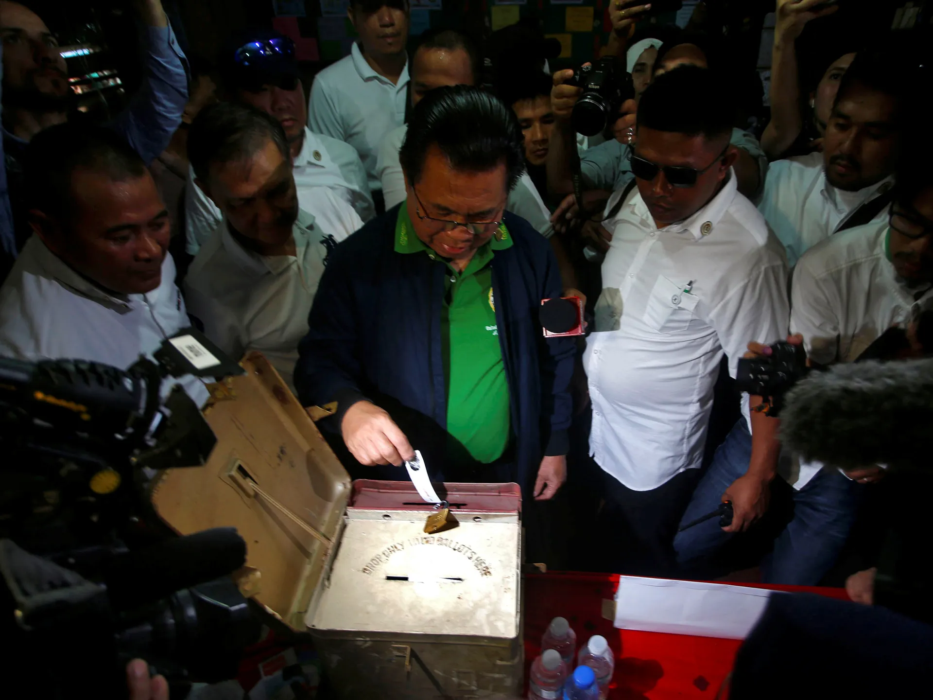 Officials and journalists crowd around a ballot box as a ballot is deposited.