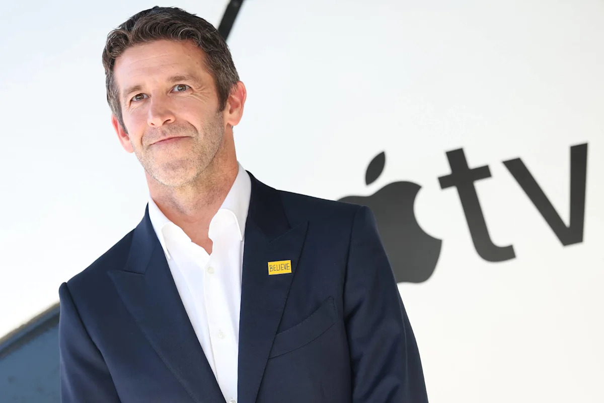 Man in navy suit wearing a yellow Believe pin in front of an Apple logo