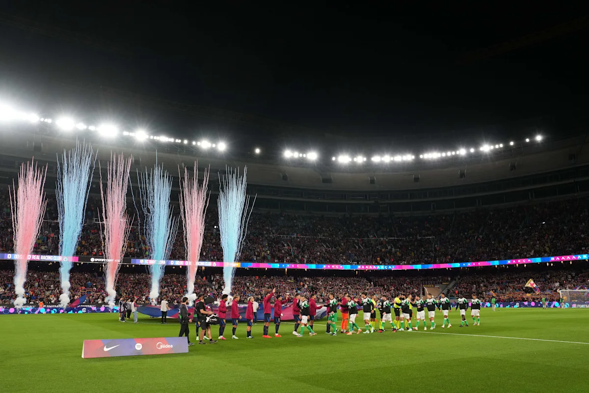Football players walk onto a brightly lit pitch as pink and blue fireworks rise before cheering spectators.