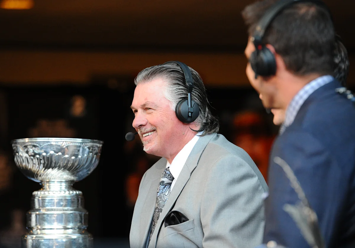 Smiling gray-haired man wearing a headset beside a large silver hockey trophy during a broadcast.