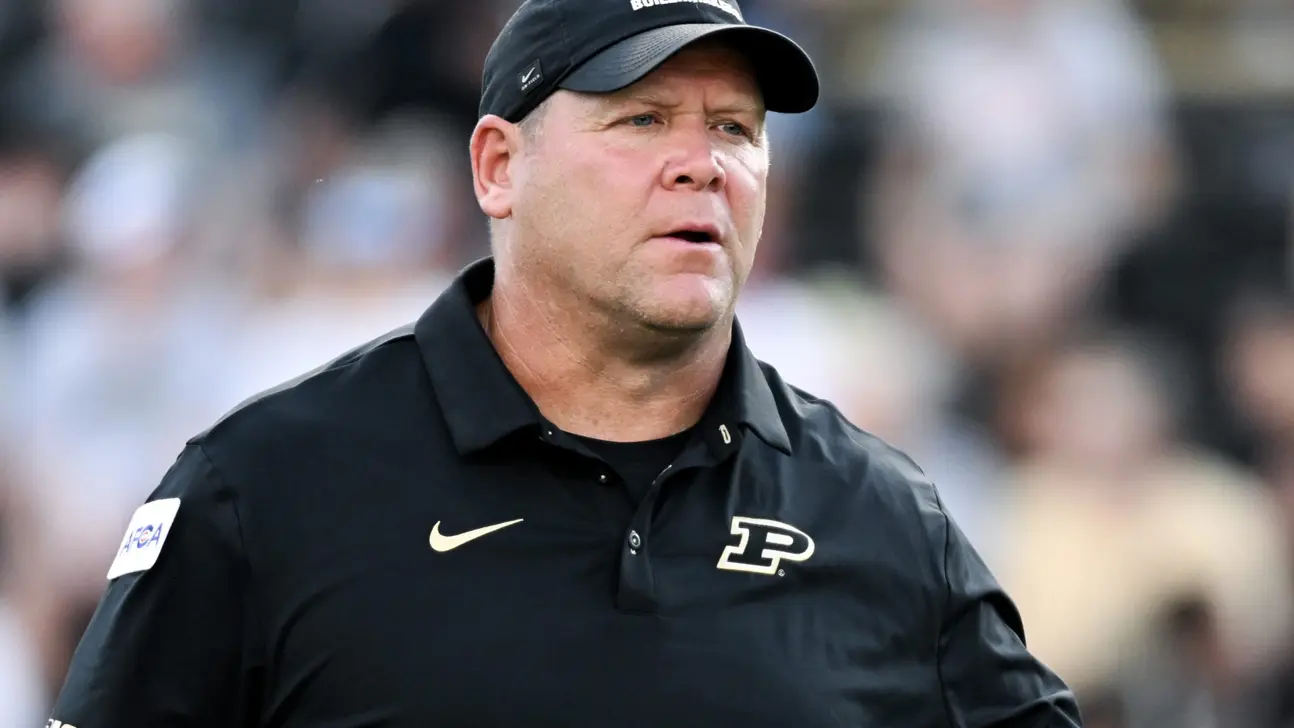 Football coach wearing a black Purdue polo and cap stands focused on the sideline.
