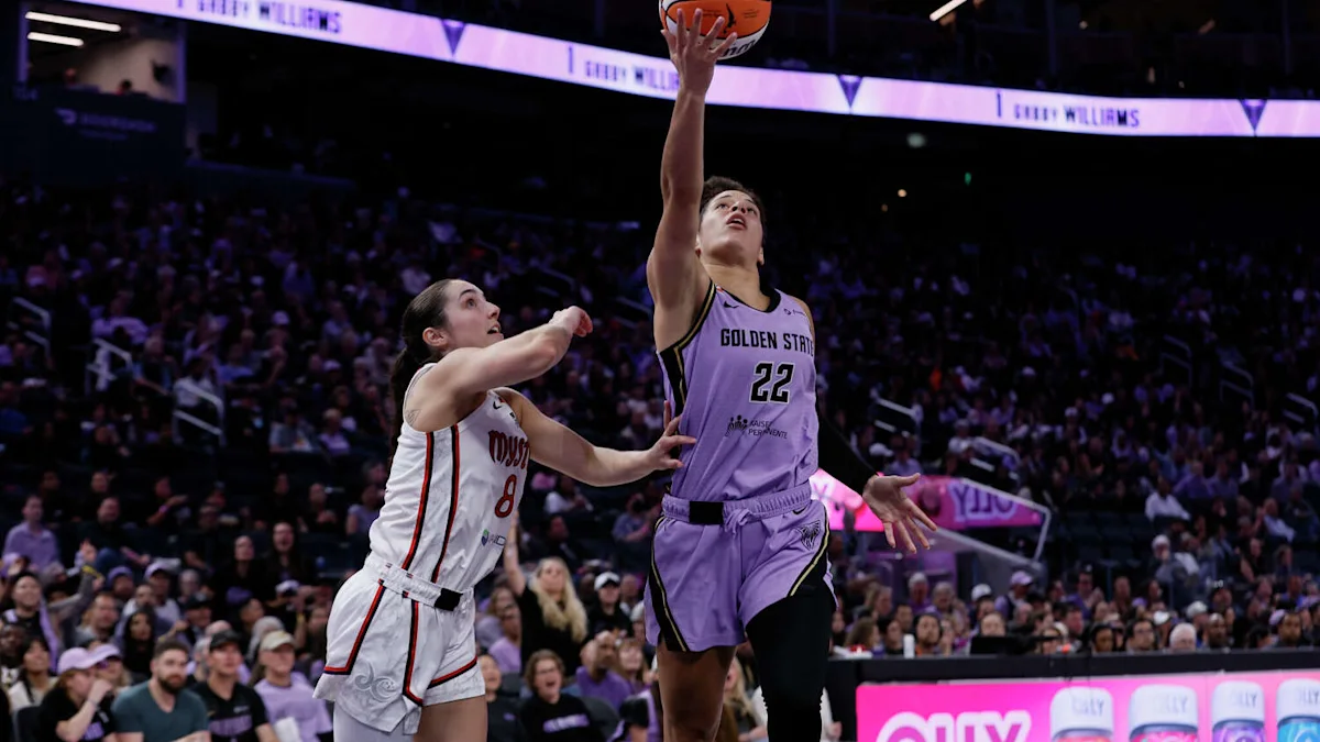 A Golden State player in purple reaches for the basket as a Dallas defender contests the shot.