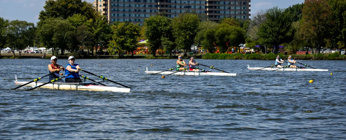 Rowers compete in pairs on a lake with trees, playground equipment, and apartment buildings behind them.