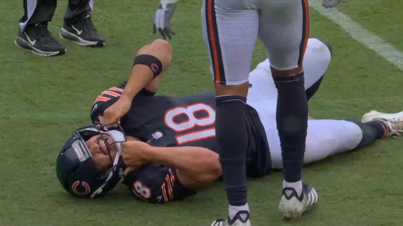 Football player in black uniform lies on field while opposing players stand nearby.