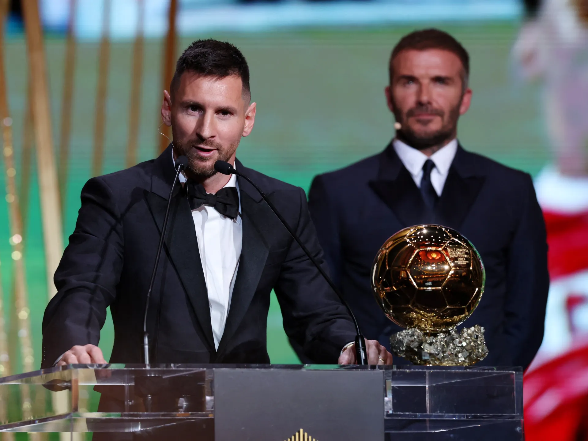 Footballer in tuxedo speaking at podium beside Ballon d’Or trophy, with another suited man behind him