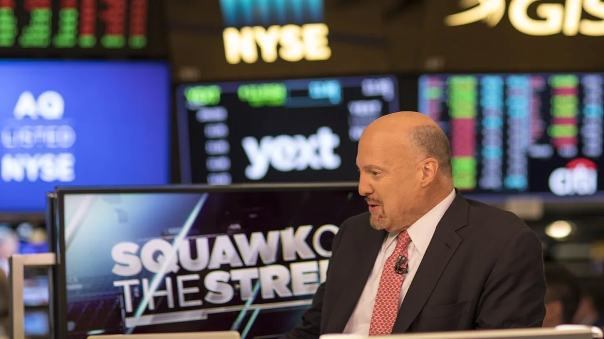 Bald man in suit and red tie speaks before stock market displays and a Squawk Box monitor