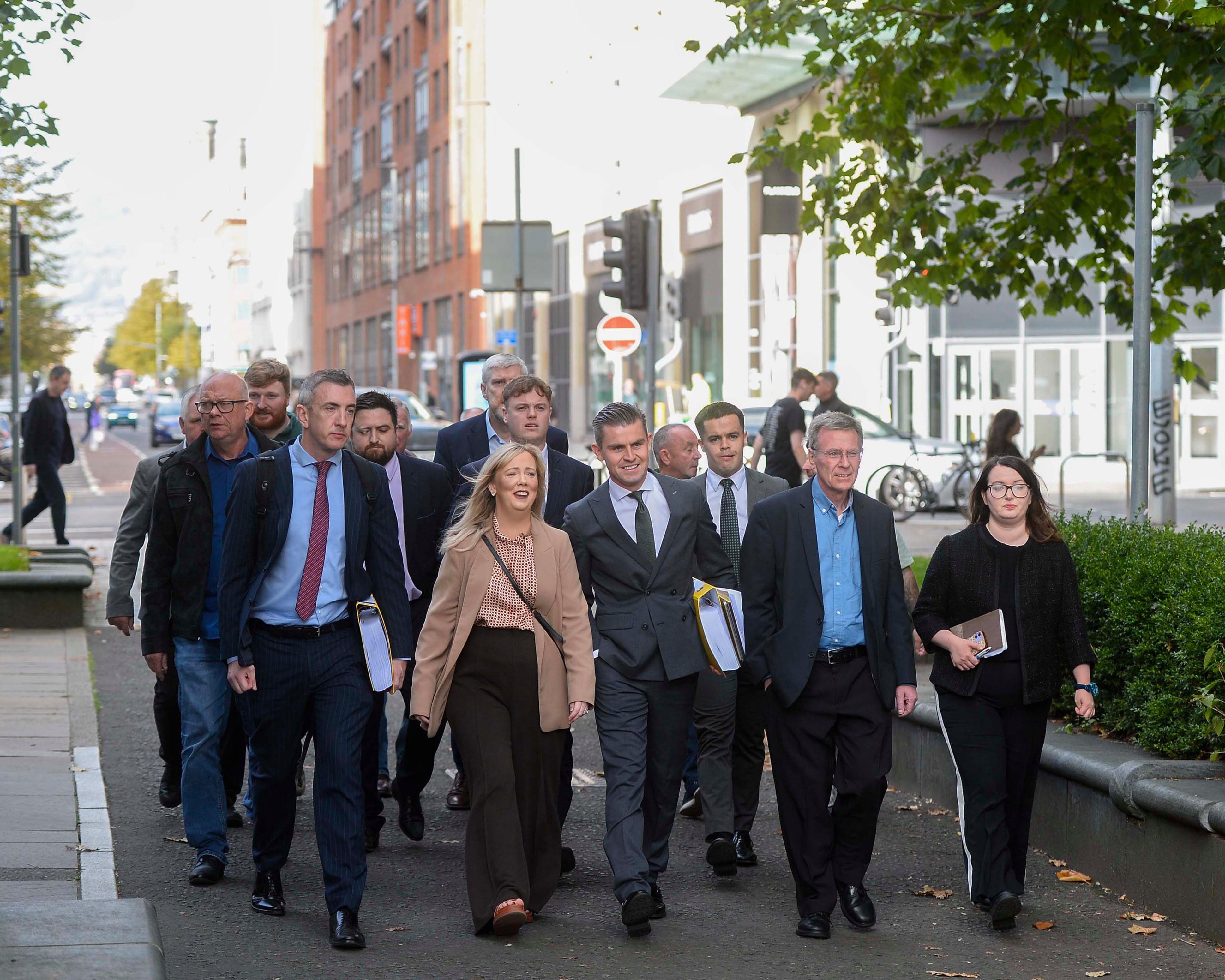 A group of formally dressed people walk along a city street beneath leafy trees.