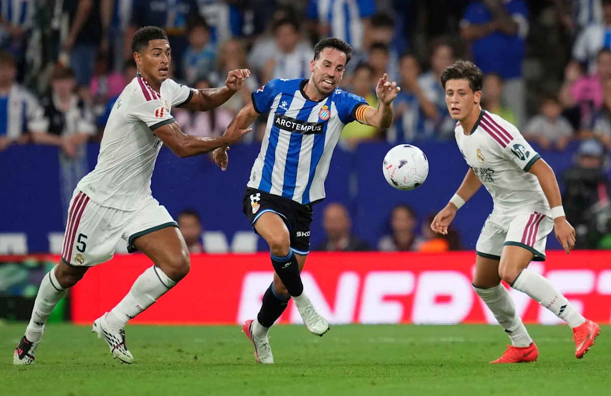 Three footballers contest the ball during a match, with two in white kits and one in blue stripes.
