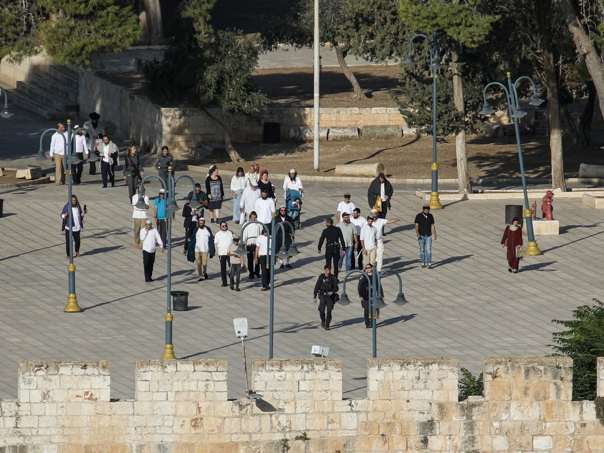 People walk across a stone-paved plaza among lamp posts, trees, and a stone wall.